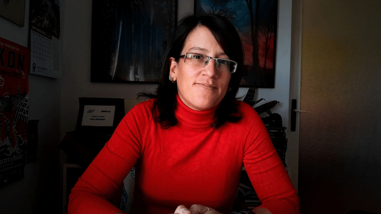 Portrati of Vesna Kurliic, a dark-haired white woman with glasses, wearing a red turtleneck, ready to present at a table, smiling slightly.